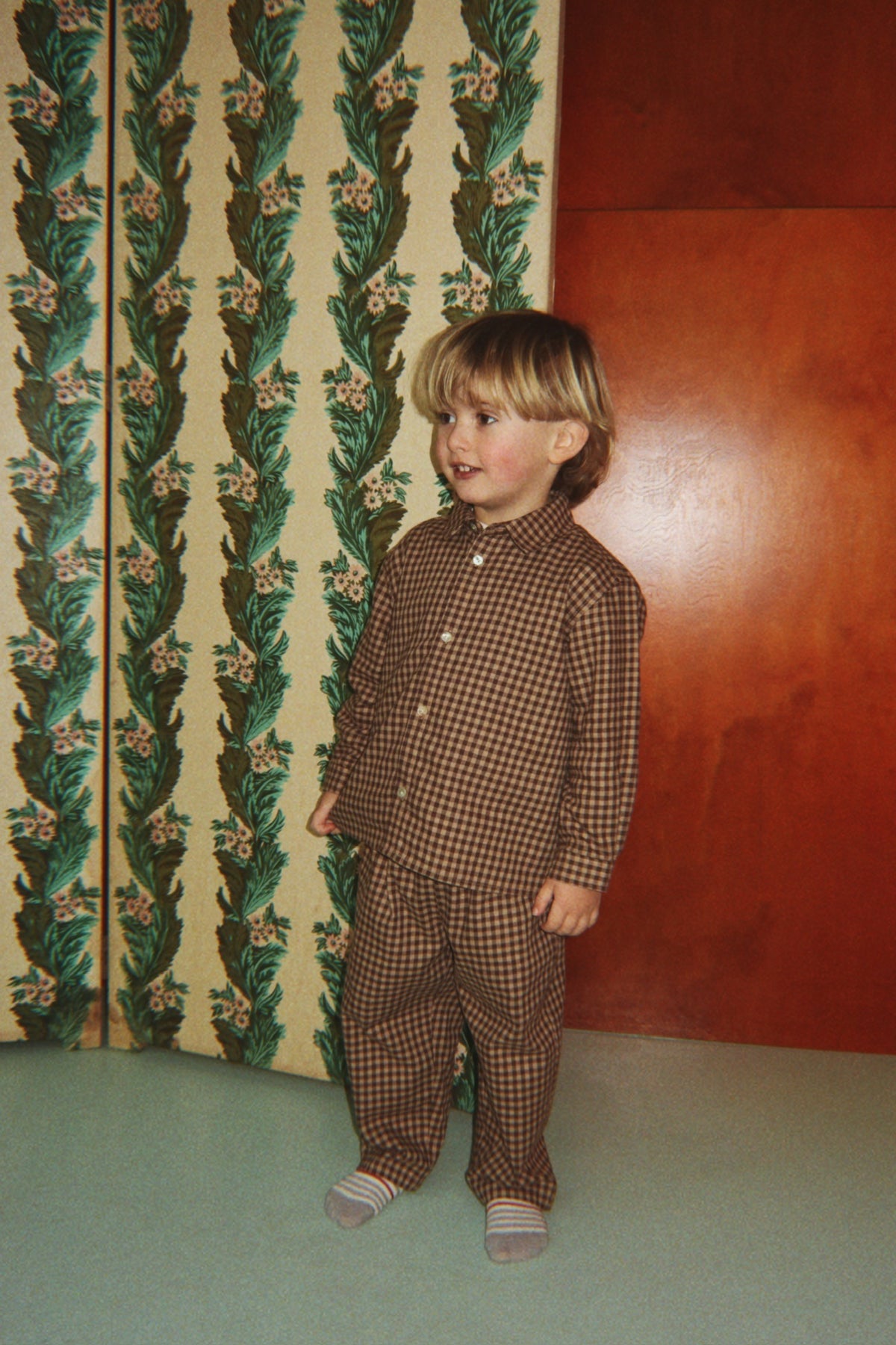 A young child wearing the Konges Sløjd Foret Long-sleeve Shirt and Pants in Chocolate Check, standing in front of a patterned green and cream folding screen and a wooden wall.