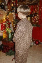 A child viewed from behind wearing the Konges Sløjd Foret Long-sleeve Shirt and Pants in Chocolate Check, standing in a decorated room with Christmas ornaments, and red wall.