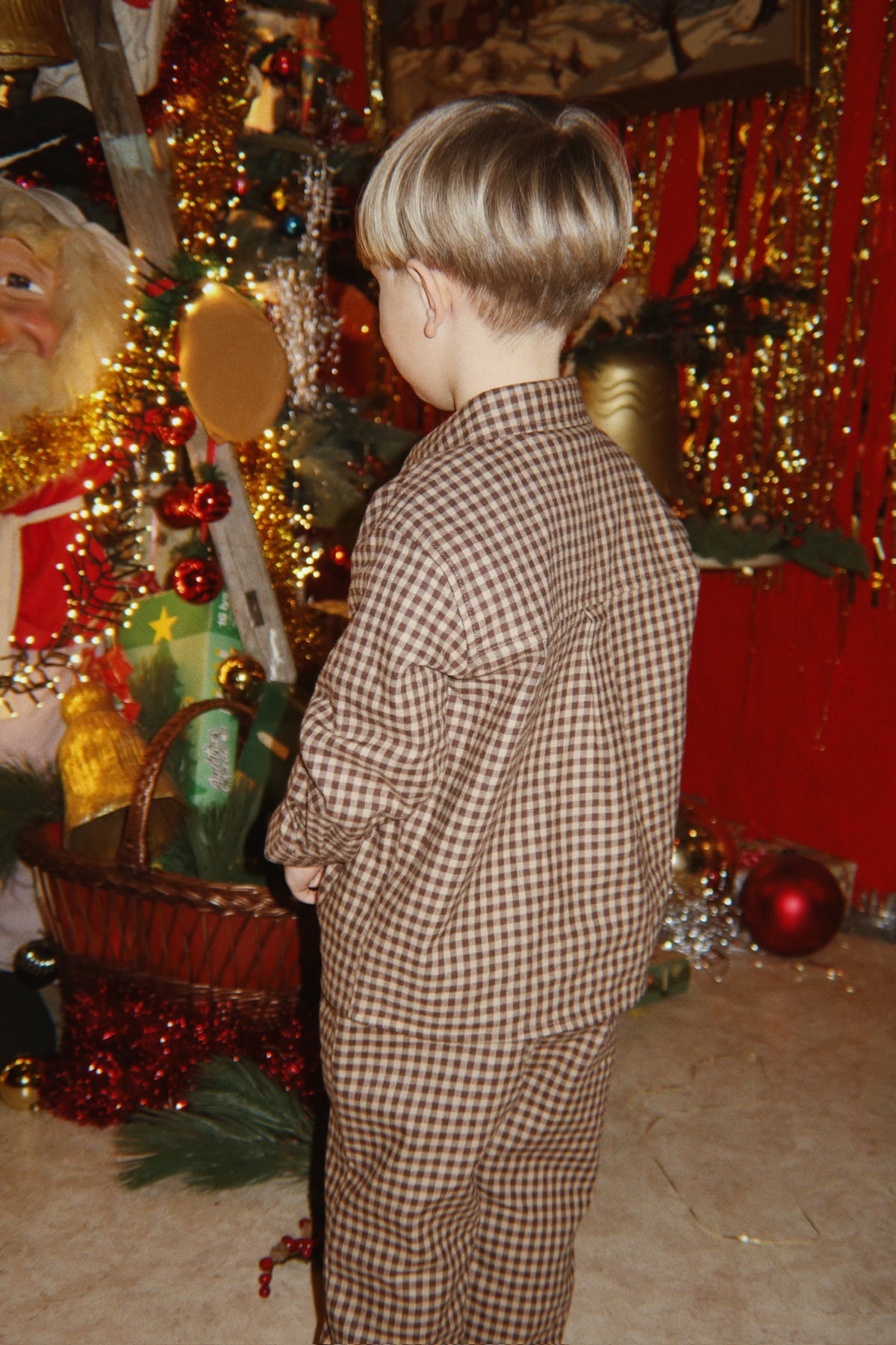 A child viewed from behind wearing the Konges Sløjd Foret Long-sleeve Shirt and Pants in Chocolate Check, standing in a decorated room with Christmas ornaments, and red wall.