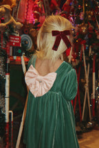 A child seen from behind wearing the Konges Sløjd Gia Bow Dress in Fir and a dark red bow in their blonde hair, standing in front of Christmas decoration.