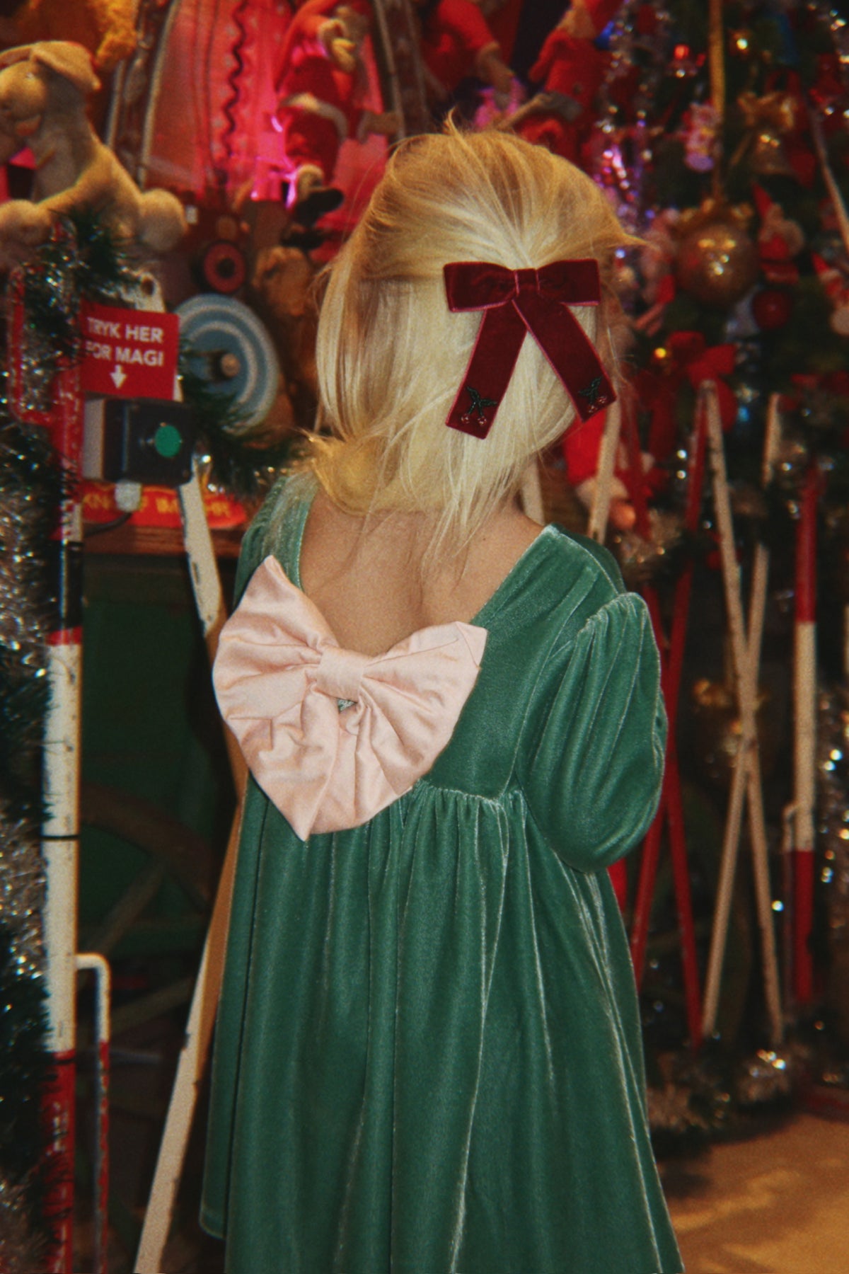 A child seen from behind wearing the Konges Sløjd Gia Bow Dress in Fir and a dark red bow in their blonde hair, standing in front of Christmas decoration.