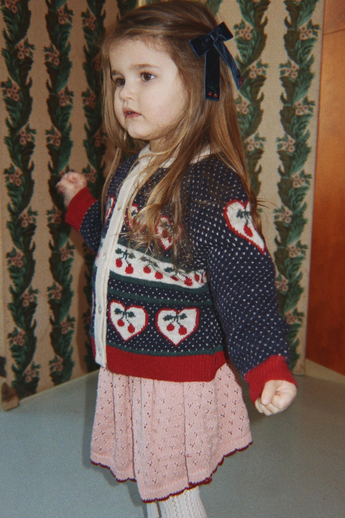 A young girl with long light brown hair and a dark blue bow in her hair, wearing a navy blue knit cardigan with red and white heart and cherry patterns (not included), a Konges Sløjd Holiday Skirt in Mellow Rose, and white socks, standing in front of a patterned green and beige room divider.