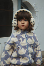 A young child wearing a Konges Sløjd Jody Teddy jacket in Swan print and earmuffs, standing outdoors in front of a white building.
