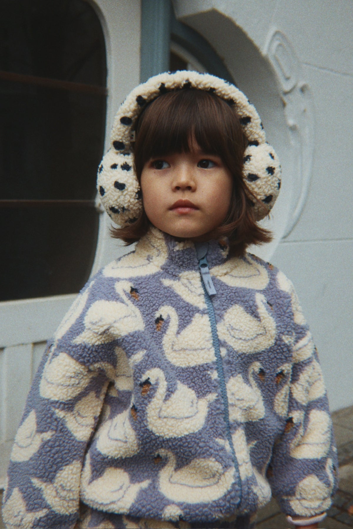 A young child wearing a Konges Sløjd Jody Teddy jacket in Swan print and earmuffs, standing outdoors in front of a white building.