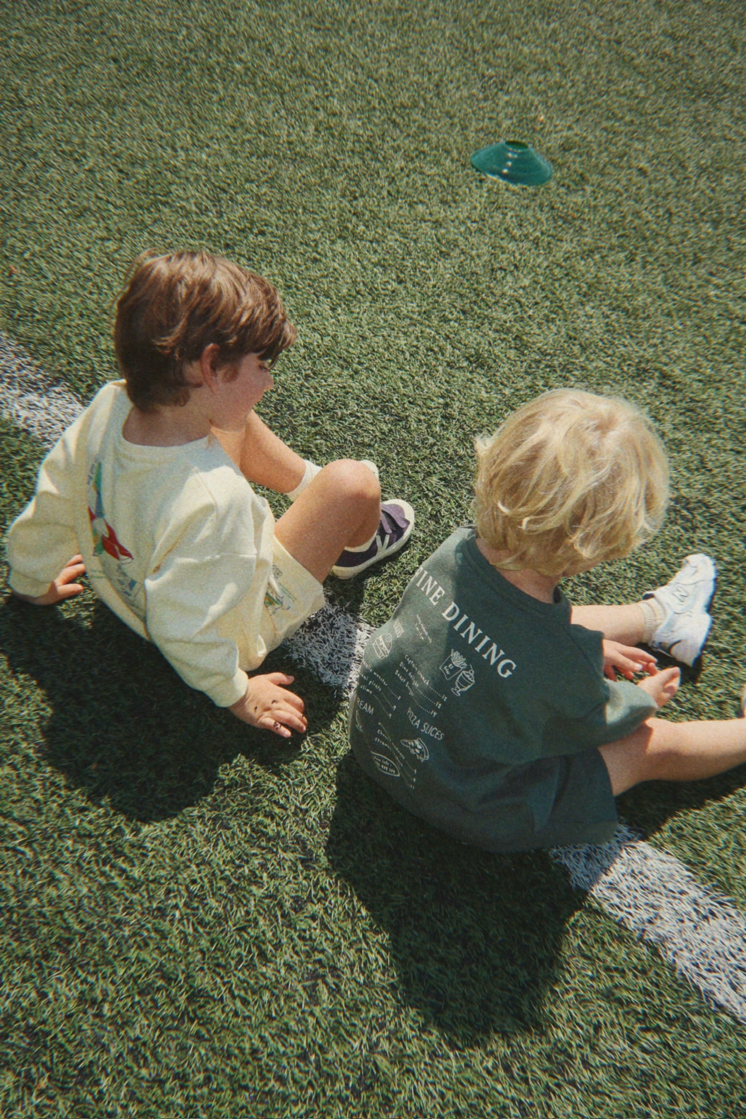 A child wearing the Konges Sløjd Lou sweat shirt in Cilantro while sitting on a soccer field with another child.