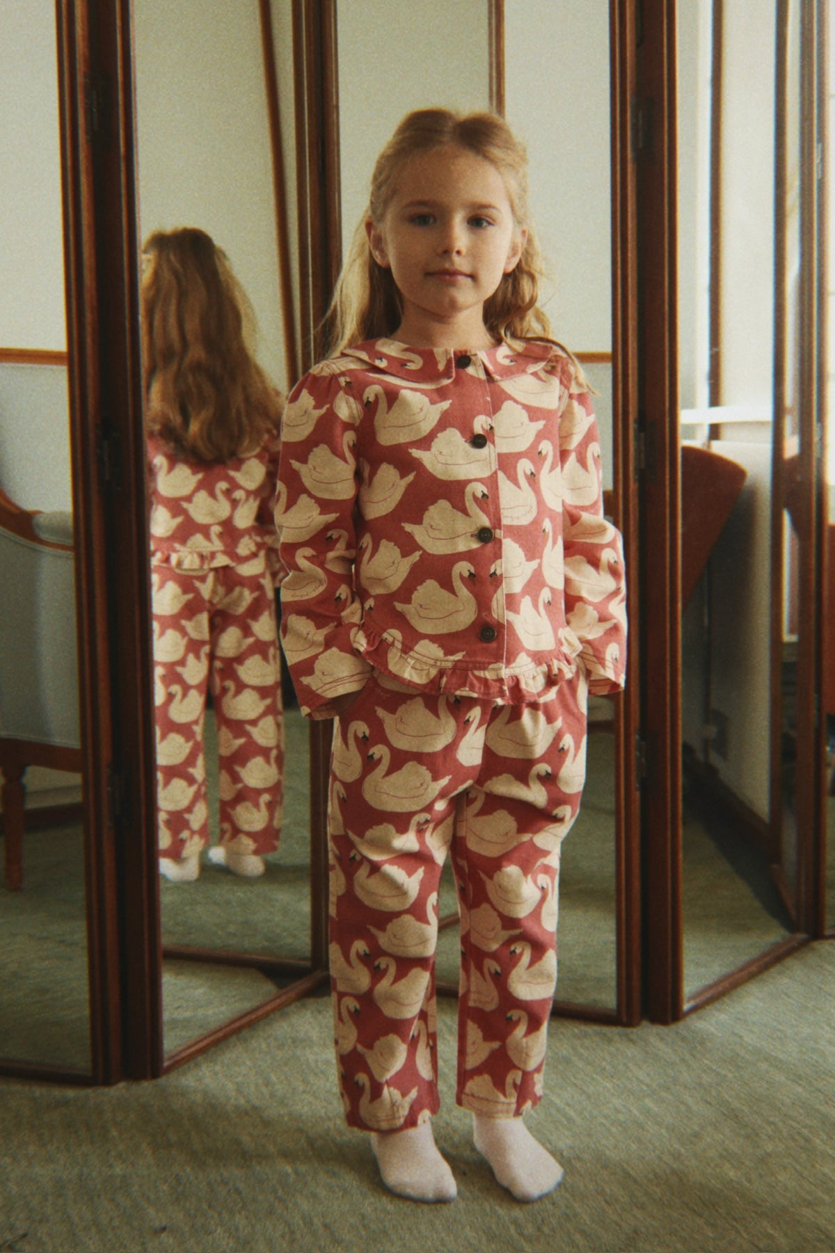 A young girl stands in front of a mirror wearing the Konges Sløjd Magot Frill Jacket in Swan Rose.