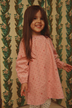 A young girl with long brown hair smiling while wearing the Konges Sløjd Marlo Dress in Cherry Dot Scarlet print, standing in front of a vintage green and beige patterned wallpaper background.