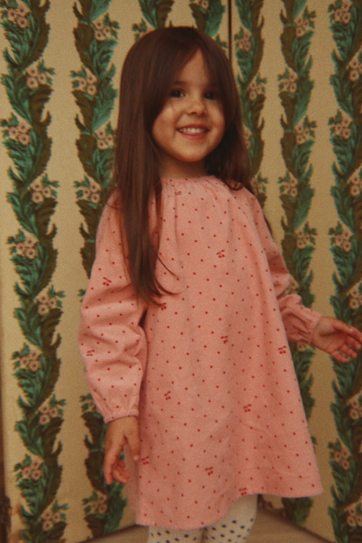 A young girl with long brown hair smiling while wearing the Konges Sløjd Marlo Dress in Cherry Dot Scarlet print, standing in front of a vintage green and beige patterned wallpaper background.