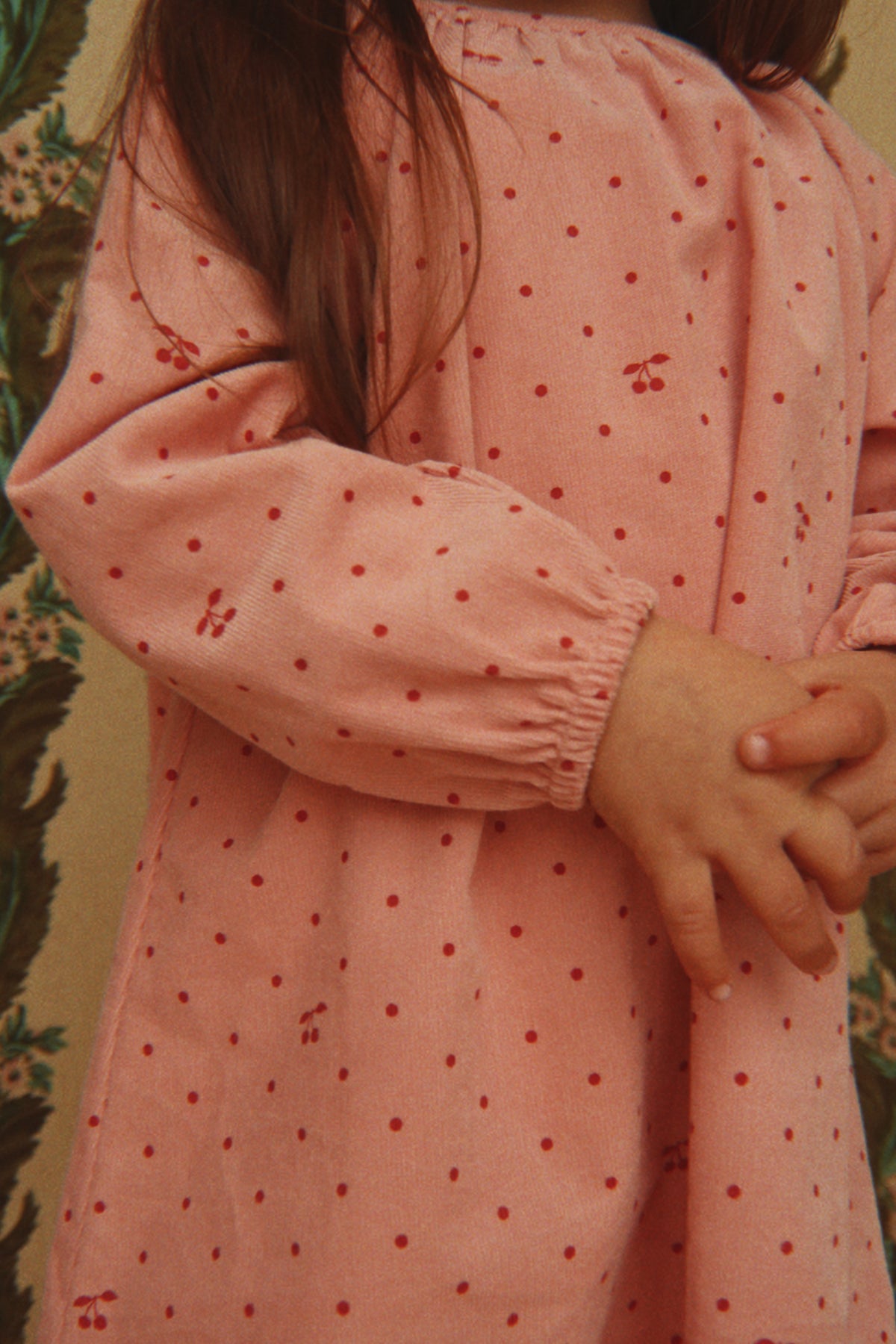 A close-up of a child wearing the Konges Sløjd Marlo Dress in Cherry Dot Scarlet print, standing against a patterned background.