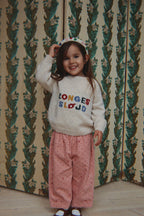 A young girl smiling and posing in front of a vintage patterned wallpaper background. She is wearing the Konges Sløjd Marlo pants in Cherry Dot Scarlet print, a white-colored knit sweater with colorful embroidered text and a white polka dot headband.