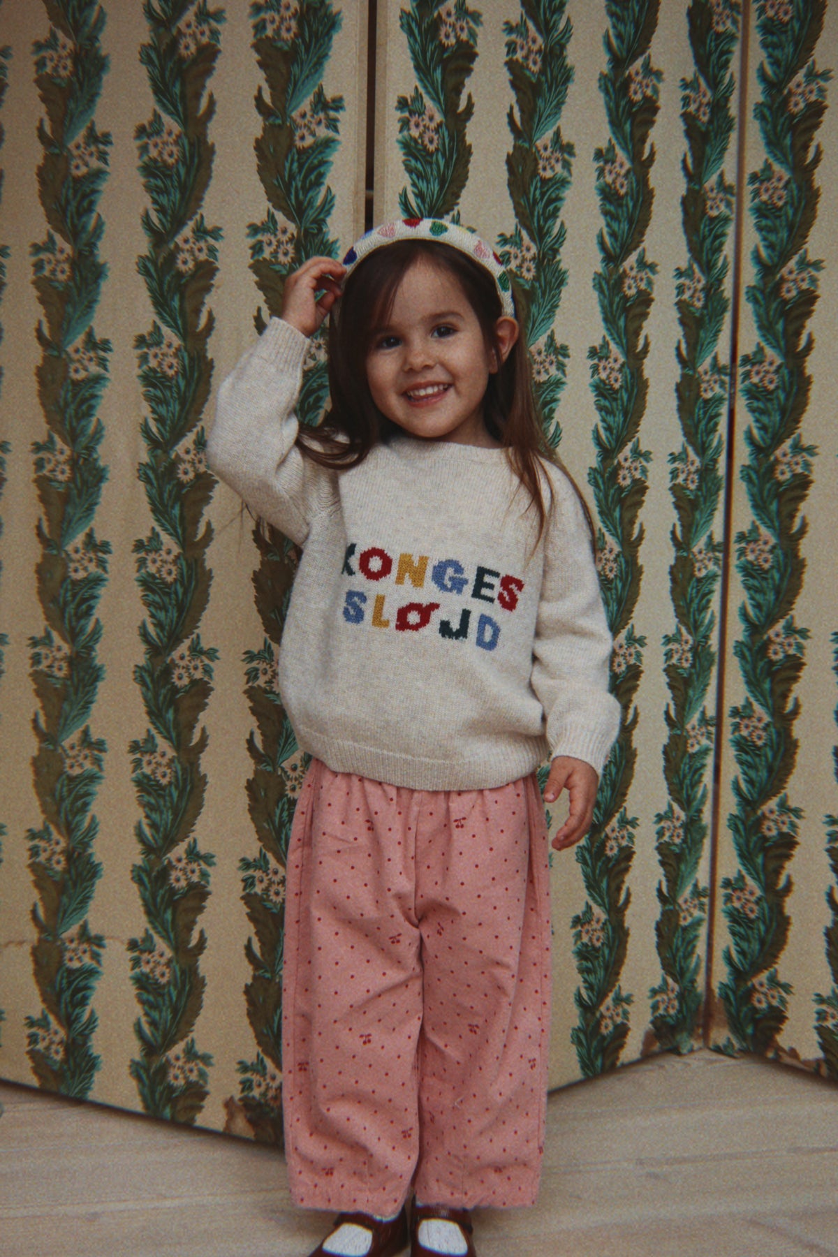 A young girl smiling and posing in front of a vintage patterned wallpaper background. She is wearing the Konges Sløjd Marlo pants in Cherry Dot Scarlet print, a white-colored knit sweater with colorful embroidered text and a white polka dot headband.