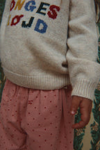 A close-up of a child wearing a cream knit sweater with colorful letters and the Konges Sløjd Marlo pants in Cherry Dot Scarlet print.