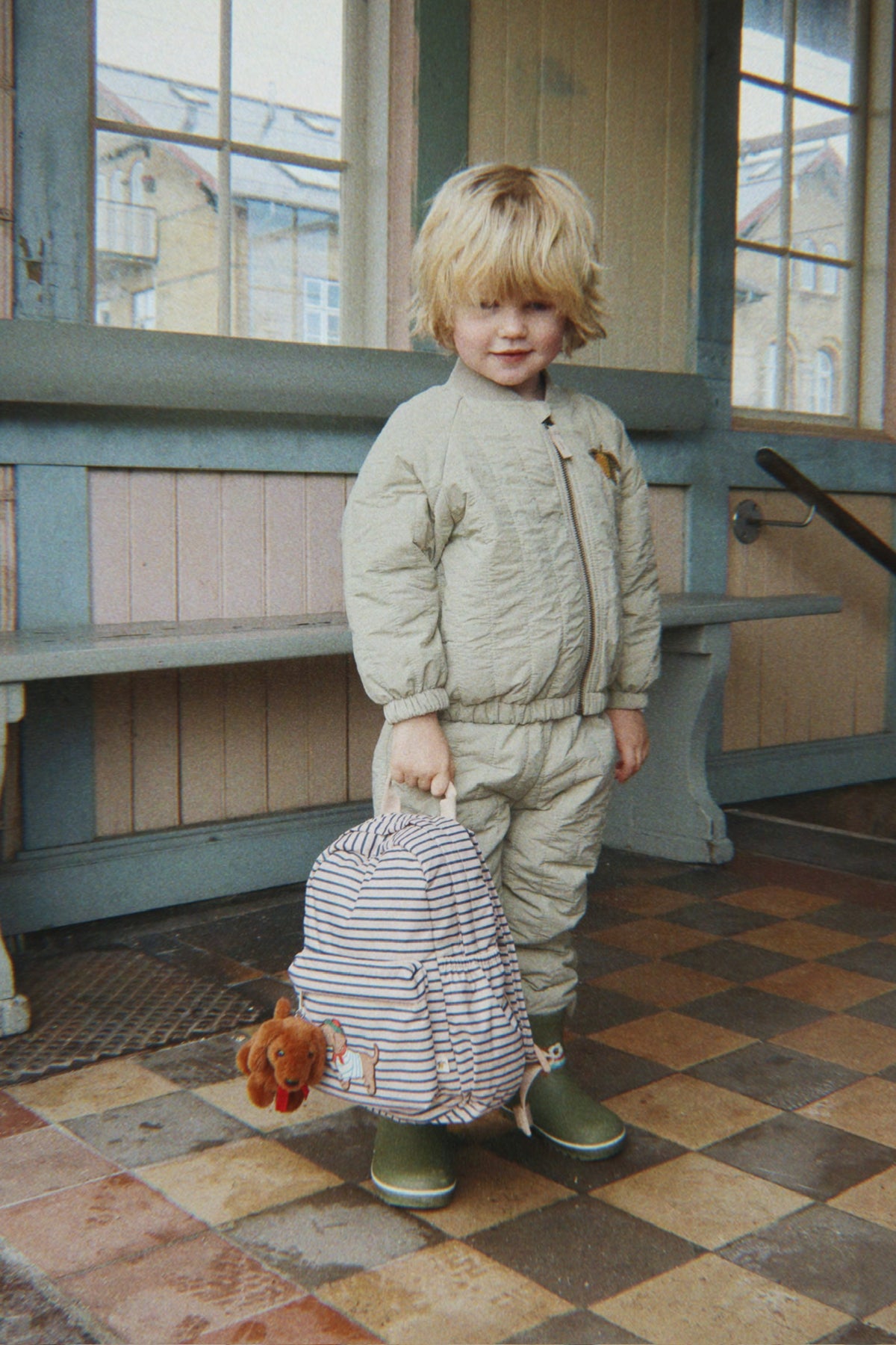 A young child wearing the Konges Sløjd Juno Terry Bomber Jacket in Laurel Oak and matching pants stands indoors holding the Nohr mini backpack in Stripe la Mer. The setting has a rustic wooden wall, windows, and a checkered floor.