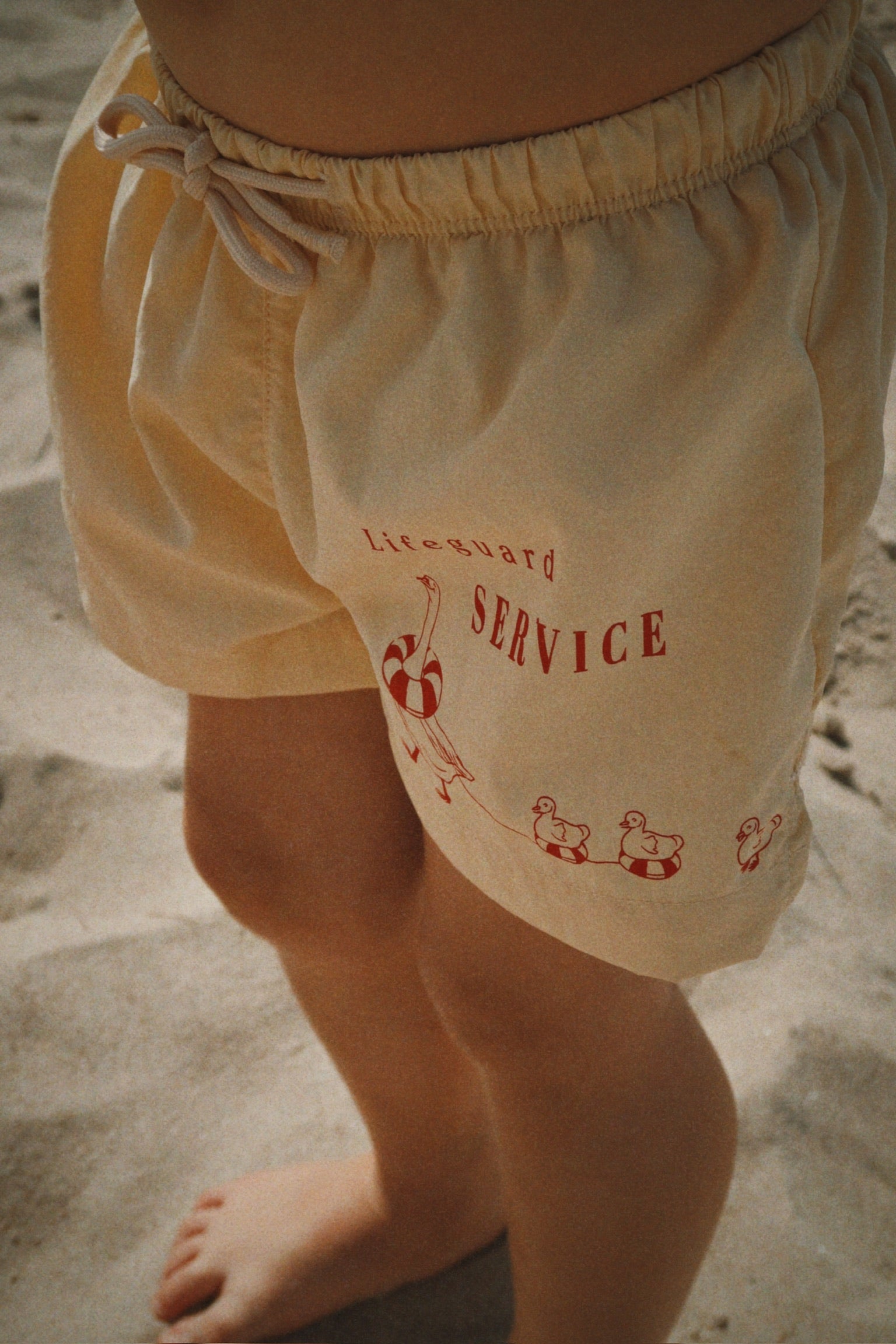 Close-up of a child wearing the Konges Sløjd Pomio short swim shorts in banana crepe on a sandy beach