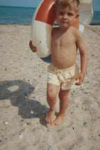 Child wearing the Konges Sløjd Pomio short swim shorts in banana crepe, while holding a life preserver on a sandy beach