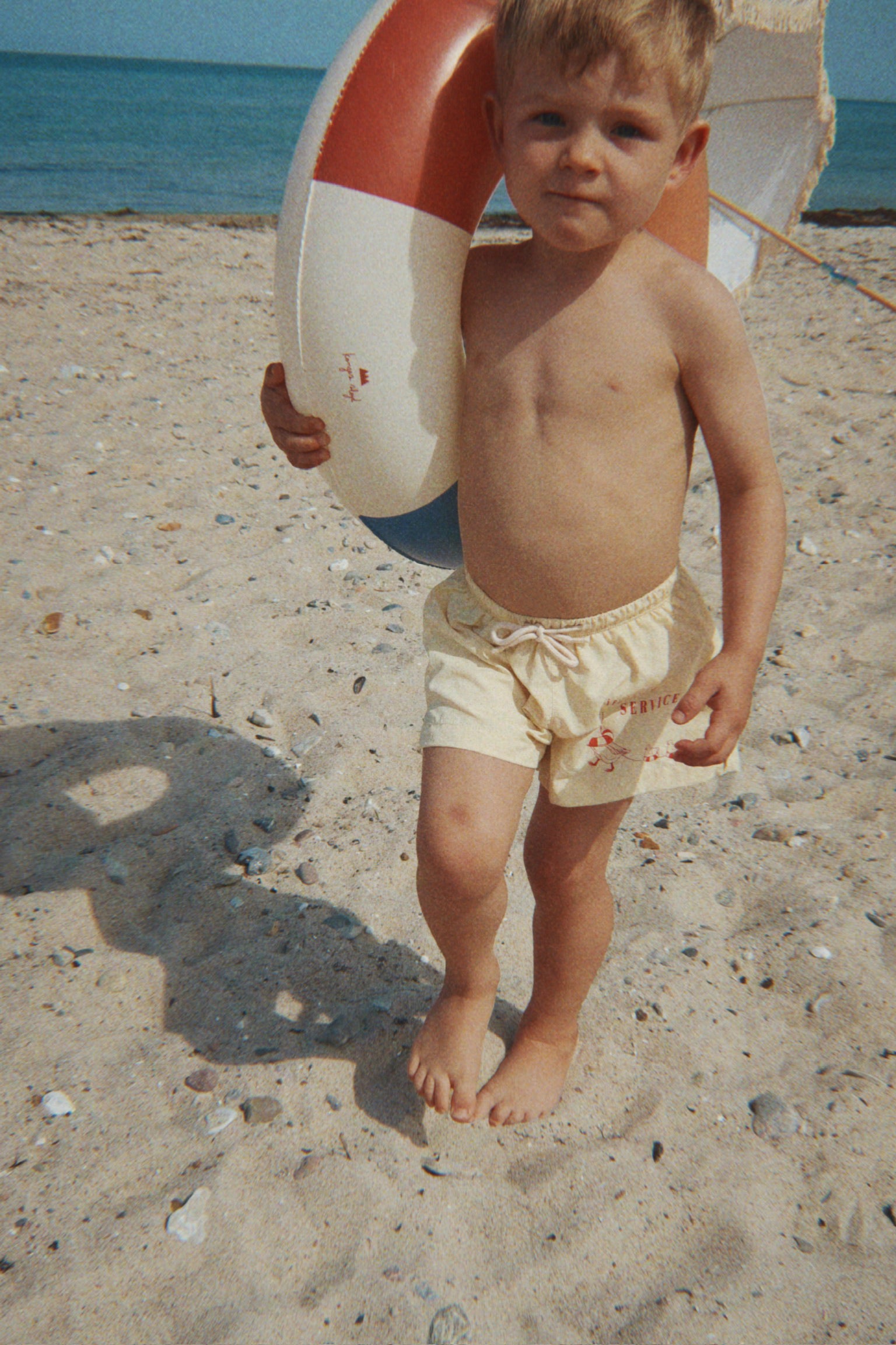Child wearing the Konges Sløjd Pomio short swim shorts in banana crepe, while holding a life preserver on a sandy beach
