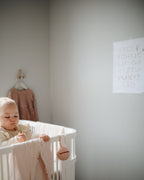 Mushie pacifier case in Natural hanging on a crib with baby in a minimalist nursery