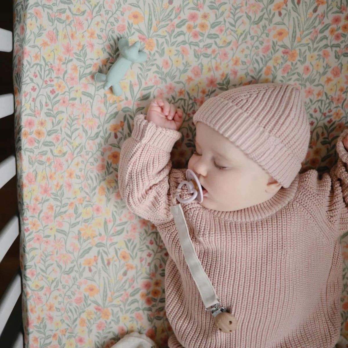 Baby in pink outfit and hat sleeping on a crib with floral bedding next to a Mushie figurine teether in Dog
