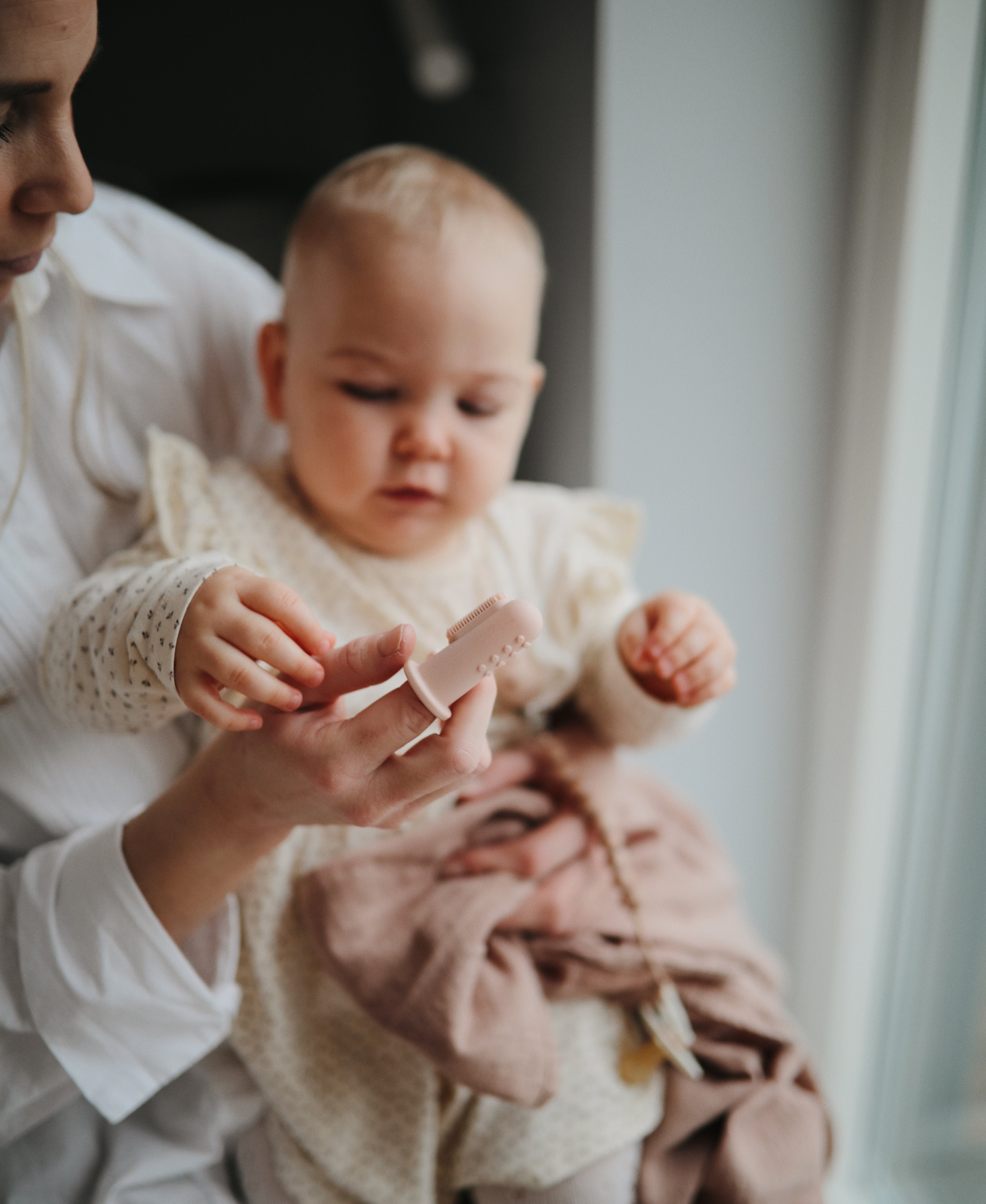 Woman holding a baby and showing them Mushie finger toothbrush Blush 