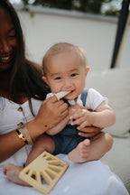 Woman brushing a baby's teeth with a Mushie finger toothbrush in Shifting sand on a blurry background