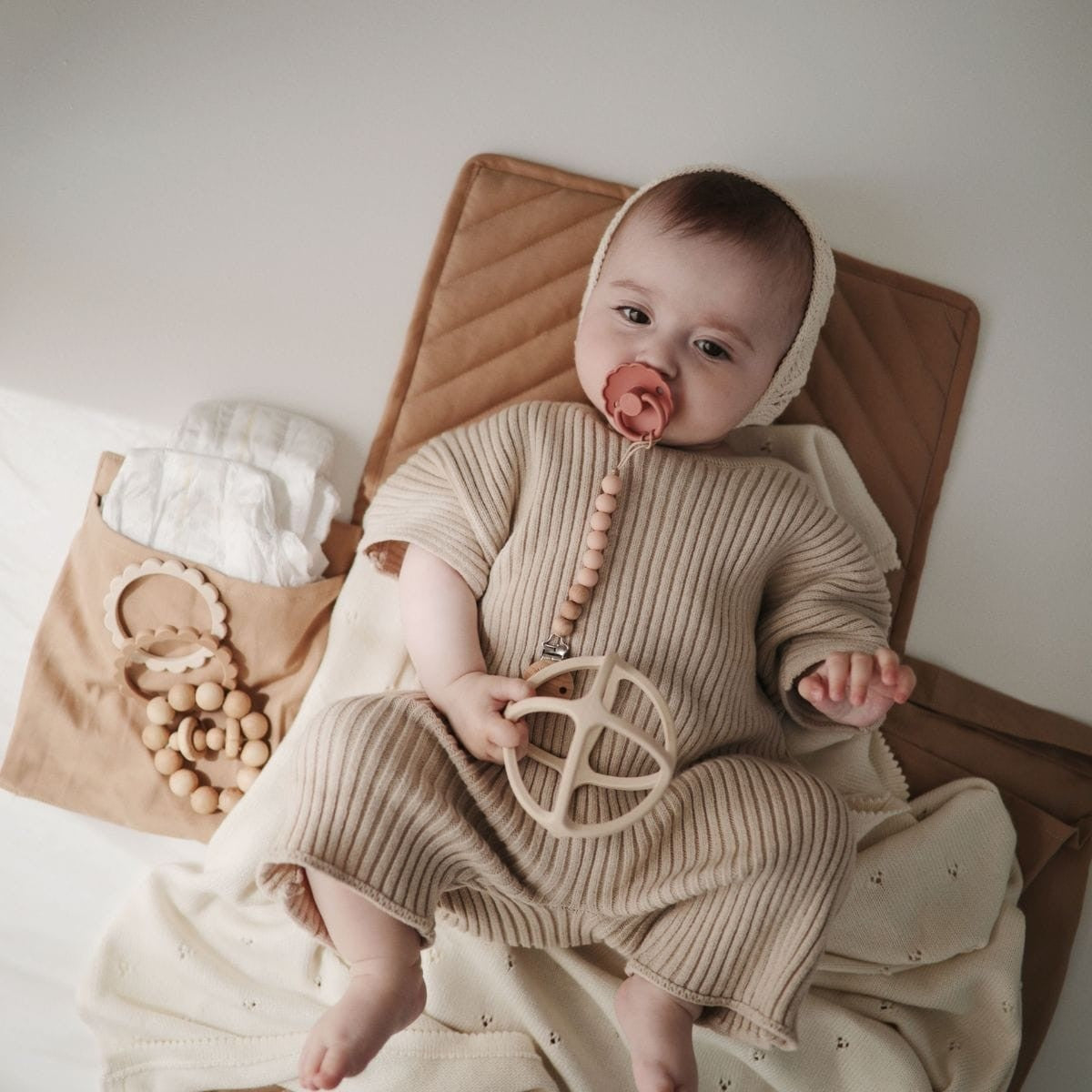 Baby in a beige outfit lying on a brown blanket styled with items from the Mushie and FRIGG Gift Box Girl