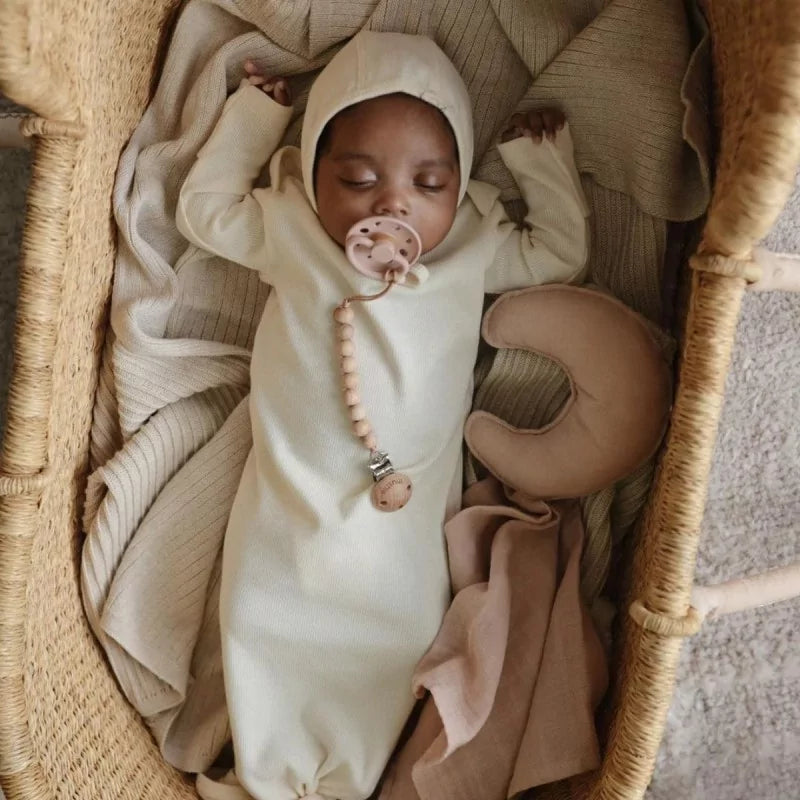 Baby in a white onesie with a pacifier, sleeping in a woven basket with a Mushie lovey blanket moon in Blush
