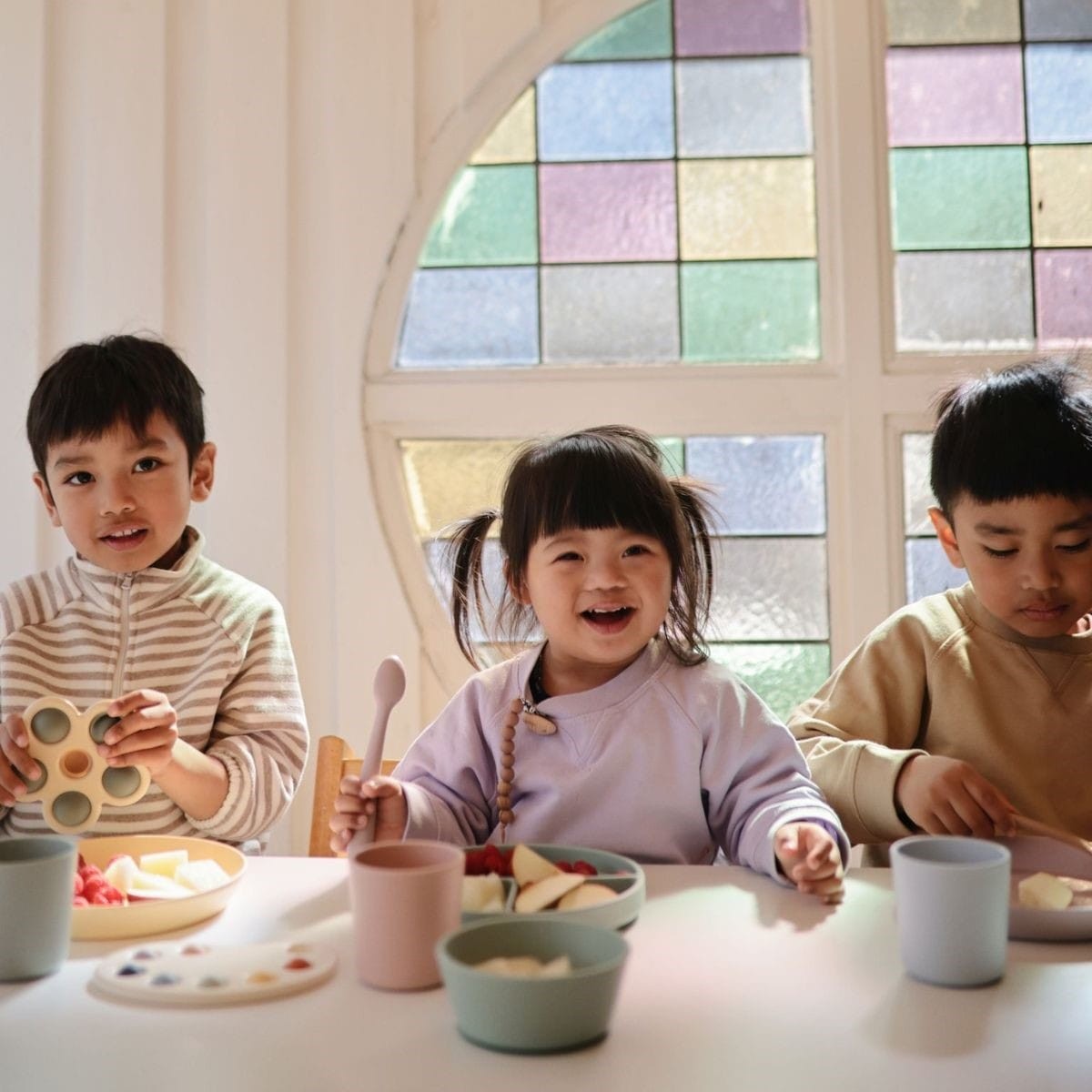Child wearing Mushie Eva pacifier clip in Cloudy Mauve sitting at a table with colorful dishes and two other children