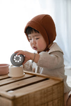 Child wearing a brown bonnet playing with Mushie stacking cups in Original