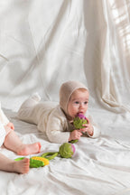 Baby wearing a beige outfit and bonnet, lying on a white blanket with Oli&Carol Artichoke baby rattle toy in natural rubber