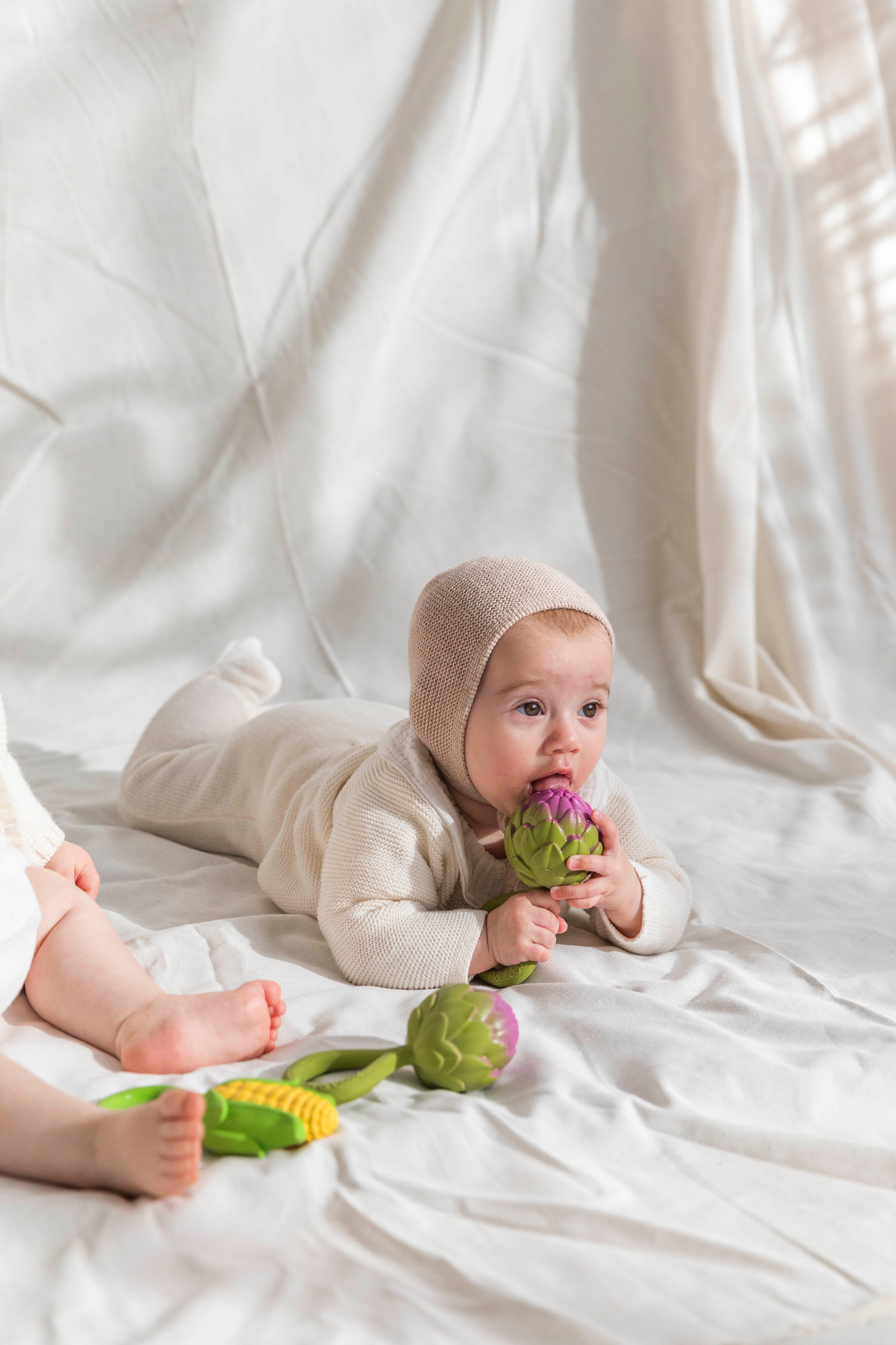 Baby wearing a beige outfit and bonnet, lying on a white blanket with Oli&Carol Artichoke baby rattle toy in natural rubber