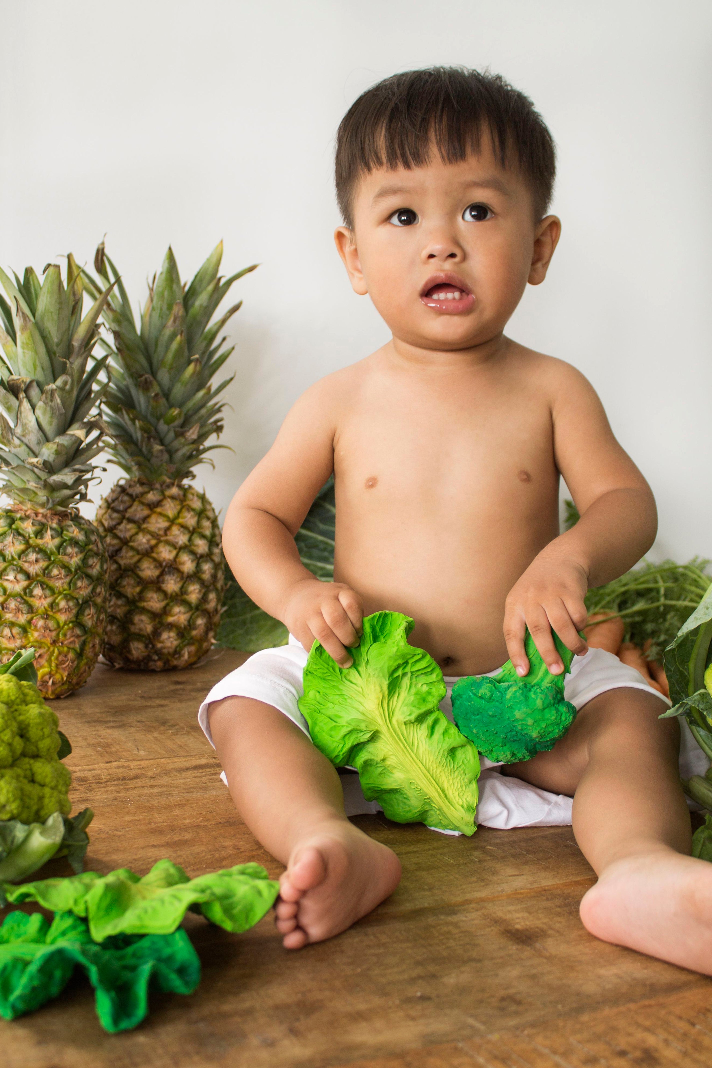Child holding Oli & Carol Kendall the Kale natural rubber teether on a wooden surface with pineapples