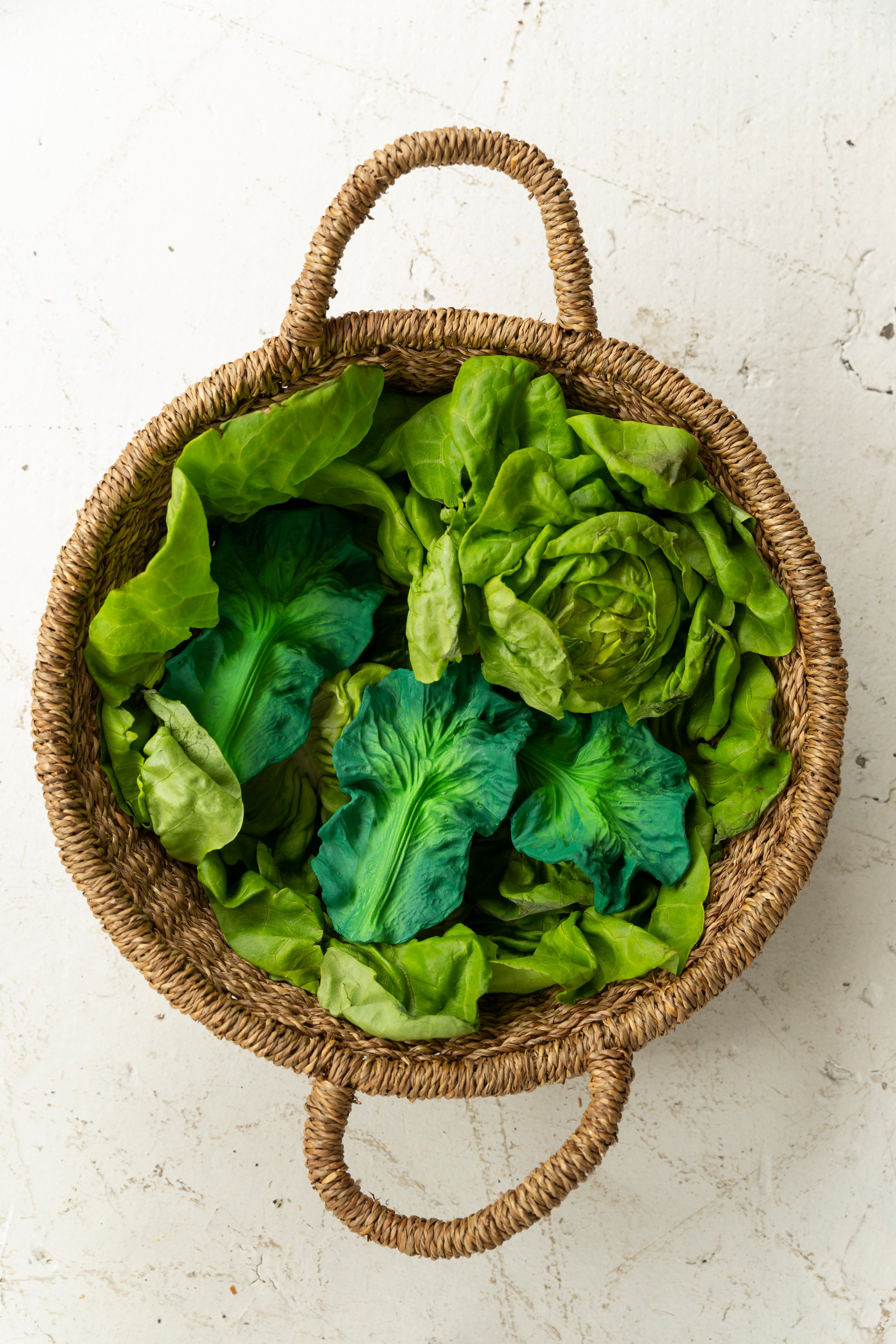 Oli & Carol Kendall the Kale natural rubber teether lying in a woven basket surrounded by green leafy vegetables on a white surface