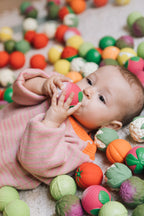 Baby holding an Oli&Carol Veggie Soup sensory ball while lying on a carpet with different sensory balls (not included)