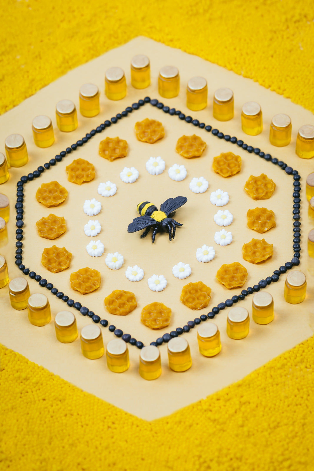Petit Boum Bees sensory bottle lifestyle image in bee theme on a yellow background showing a bee, honeycomb, honey jars and daisies arranged around it