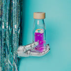 Lifestyle image of a white-gloved hand holding the Petit Boum Magic Rainbow sensory bottle as it emerges from a silver glitter curtain