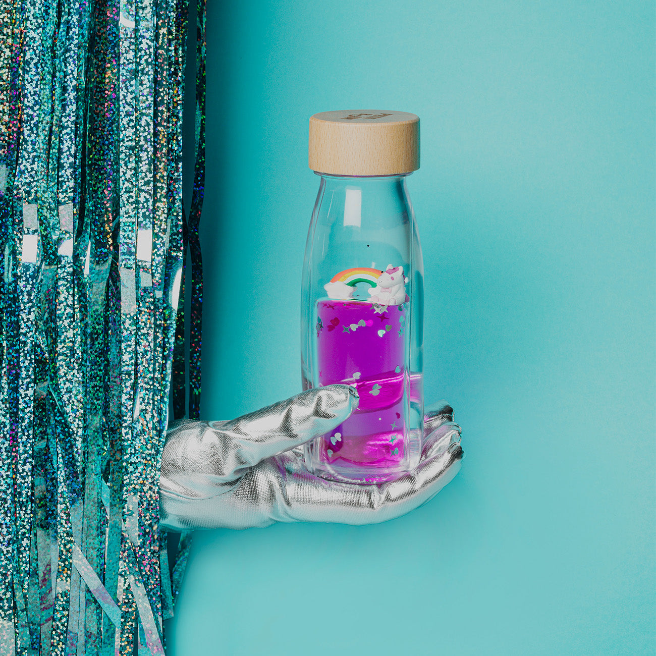 Lifestyle image of a white-gloved hand holding the Petit Boum Magic Rainbow sensory bottle as it emerges from a silver glitter curtain