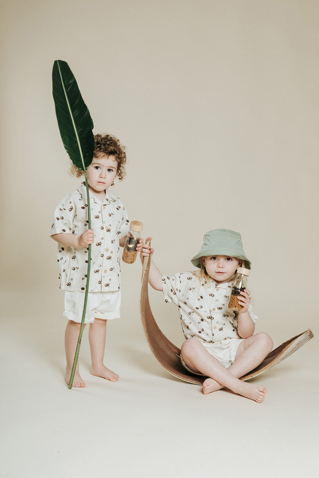 Petit Boum Monkey sensory bottle lifestyle image with two children in matching outfits holding the sensory toy, styled with a leaf and wooden scooter on a beige background