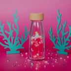 Lifestyle image of a Petit Boum Sea sparkle sensory bottle, surrounded by teal coral, decorative elements and against a pink background