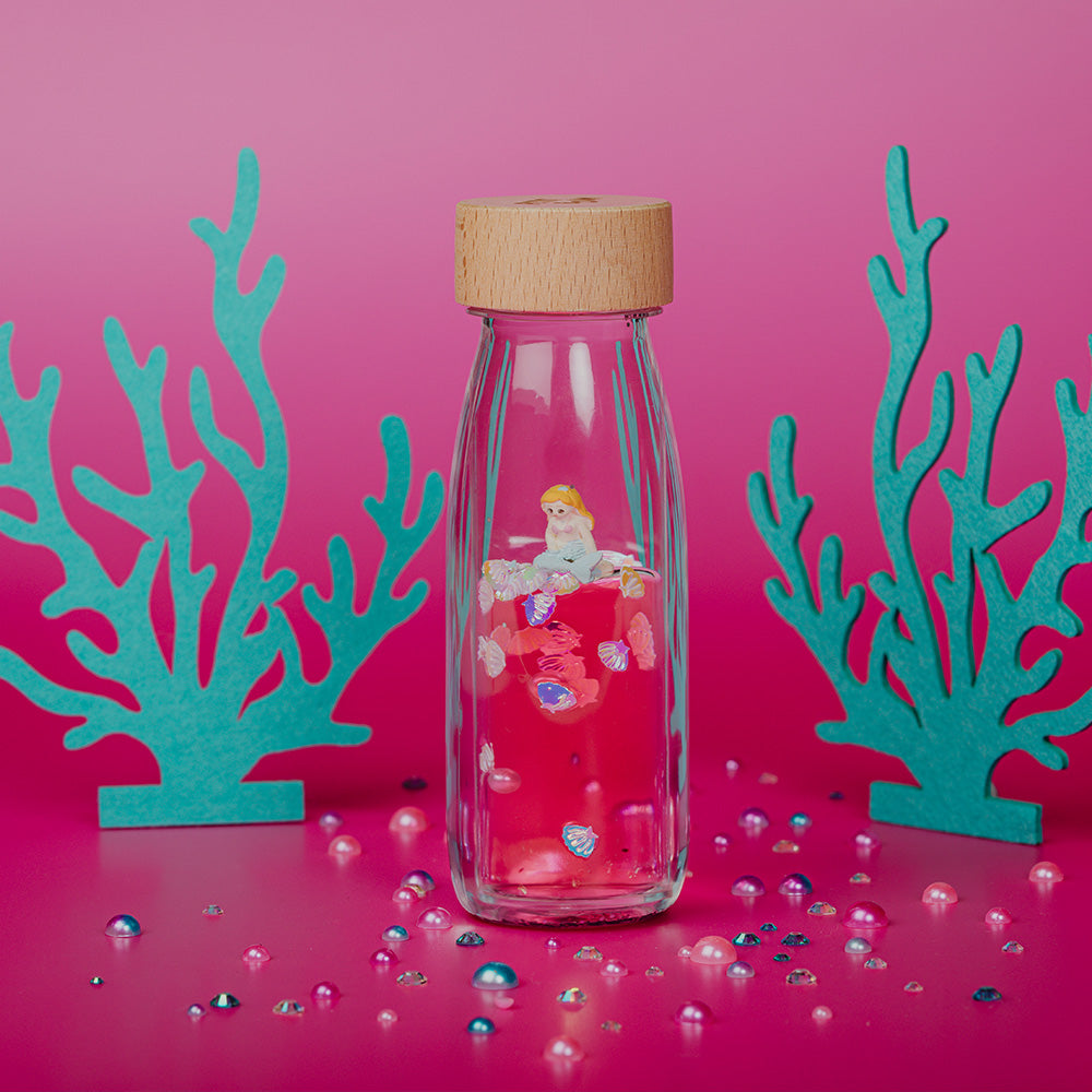 Lifestyle image of a Petit Boum Sea sparkle sensory bottle, surrounded by teal coral, decorative elements and against a pink background