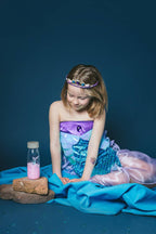 Lifestyle image of a child in a mermaid costume sitting on the ground and looking at one of the Petit Boum Fantasy set of 3 sensory bottles 