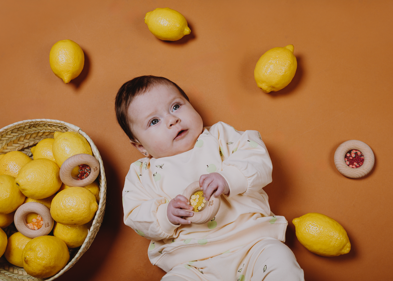 
Petit Boum Donut Lemons lifestyle image of a baby holding the bottle against an orange background surrounded by lemons and a lemon basket