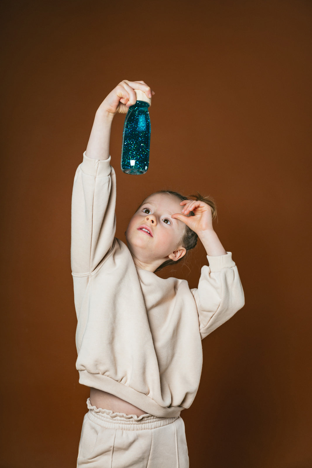 Lifestyle image of a girl holding the Petit Boum Turquoise sensory bottle up and looking at it from below