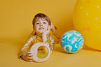 Lifestyle image of a child in a gold outfit holding the Petit Boum sensory Moon, surrounded by a planet Earth ball and a large yellow ball on a yellow background