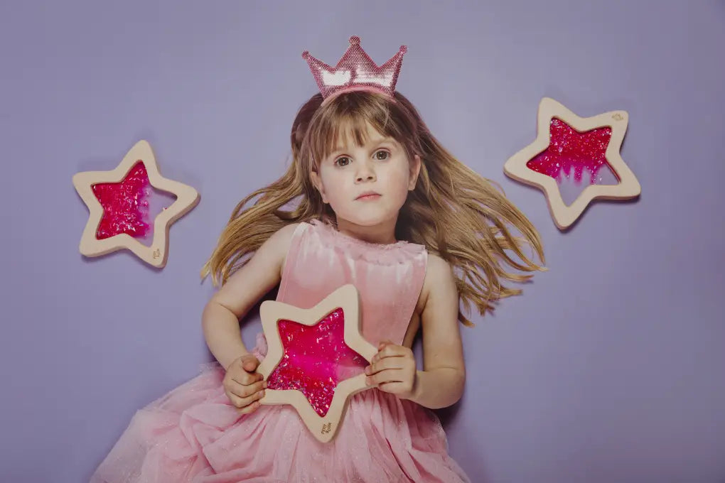 Petit Boum sensory star Nébula lifestyle image of a girl in a pink dress and pink crown lying on the floor holding the bottle with two sensory stars nearby on a purple background