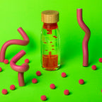 Petit Boum Spiral Pink sensory bottle styled with pink curved objects and red play balls on a green background